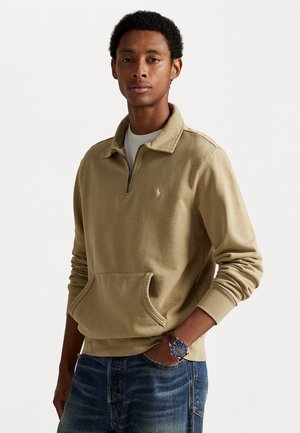 Beige kwart-rits sweatshirt met een kraag, voorzak en klein logo. Draagt bij blauwe spijkerbroeken en een horloge, met een gestructureerde stof.