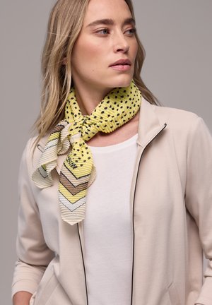 Femme aux cheveux blonds portant une veste beige clair zippée par-dessus un haut blanc et une écharpe jaune à motifs nouée autour du cou.