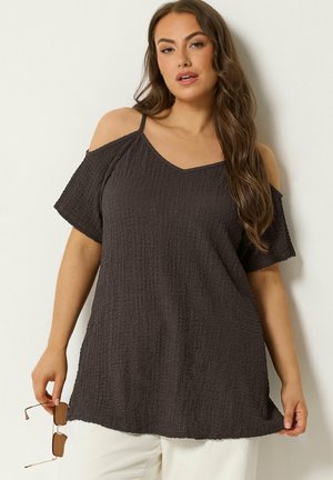 Kvinde med langt brunt hår iført en tekstureret sort cold-shoulder top og holder solbriller i den ene hånd mod en ensartet lys baggrund.
