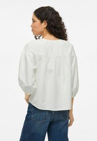 Lys grå bluse med puffermer og pyntet bakseam. Mykt stoff, avslappet passform og rund hals. Kombinert med blå denim bukser.