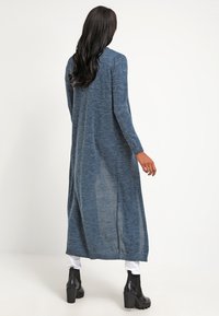 Cardigan lungo in maglia di colore blu scuro con una vestibilità ampia, maniche lunghe e una texture leggermente trasparente. Presenta dettagli di cucitura sottili.