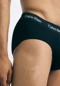 Čierne bavlnené nohavičky s jemným elastickým pásom, na ktorom sa nachádza logo "Calvin Klein" v bielej farbe. Hladká textúra a priliehavý strih zvýrazňujú postavu.