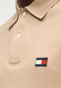 Μπεζ πόλο πουκάμισο με ραβδωτή υφή, κούμπωμα με τρία κουμπιά και κεντημένο λογότυπο της μάρκας σε κόκκινες, λευκές και μπλε αποχρώσεις στο στήθος.