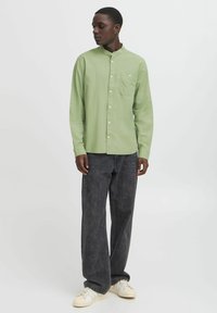 Camicia a maniche lunghe verde chiaro con colletto rialzato e taschino singolo sul petto, abbinata a pantaloni ampi grigio scuro e sneaker color crema.
