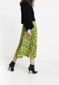 Donna che cammina indossando un maglione nero, una gonna midi con stampa serpente verde e stivaletti neri con tacco, su uno sfondo bianco.