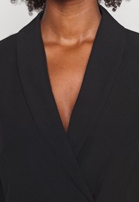 Svart blazer med en djup V-ringning, slät tyg och strukturerade slag. Närbild visar textur och rena linjer runt krageområdet.