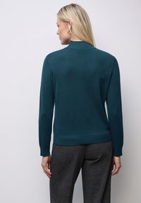 Teal trui met hoge hals en lange mouwen, gemaakt van een gladde gebreide stof, gecombineerd met grijze broeken.