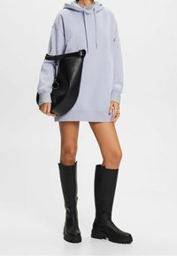 Fialové mikinové šaty s texturovaným, oversized designem, kombinované s vysokými černými koženými botami se zipem a elegantní černou kabelkou přes rameno.