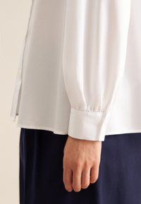Camicia bianca a maniche lunghe con polsini aderenti, texture liscia e dettagli con spacchi laterali, indossata con pantaloni blu scuro.