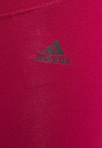 Dopasowane różowe legginsy sportowe wykonane z gładkiego, elastycznego materiału, z czarnym logo Adidas na dolnej lewej stronie.