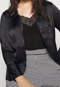 Blusa di raso nera con maniche lunghe e bottoni frontali sopra una maglietta nera con pizzo, abbinata a una gonna a scacchi in bianco e nero.