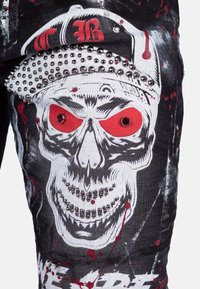 Pantaloni in denim neri con una grande grafica di teschio con accenti rossi. Borchie argentate lungo la parte superiore. Schizzi di vernice aggiungono dettagli.