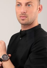 Orologio nero con quadrante rotondo, dettagli dorati, cinturino nero e funzionalità cronografiche. Indossato su un polso, abbinato a una camicia nera a bottoni.