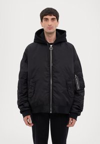 Giubbotto bomber imbottito nero con chiusura frontale a zip, cappuccio e tasche laterali, con etichetta sulla manica sinistra. Tessuto liscio e lucido.