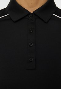 Schwarzes Polohemd mit Kragen, vier schwarzen Knöpfen und weißen Paspelierungen an den Schultern, getragen auf der oberen Brust und dem Hals einer Person.