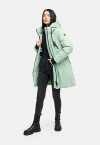 Mintgrøn isoleret parka med hætte, lynlås foran og sorte detaljer. Båret over en sort bluse og parret med sorte jeans og støvler.