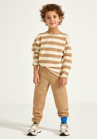 Beige Langarmhemd mit braunen horizontalen Streifen, kombiniert mit beigen Jogginghosen mit strukturierten Knieakzenten und schwarzen, weißen und grauen Sneakers.