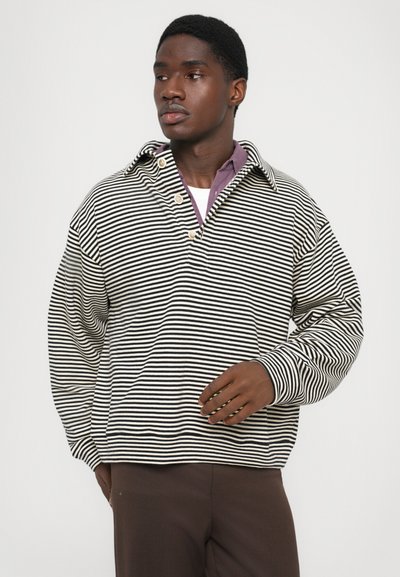 Un homme portant une chemise à rayures noires et blanches à manches longues superposée à une chemise violette à col et un pantalon marron, regardant vers sa gauche.