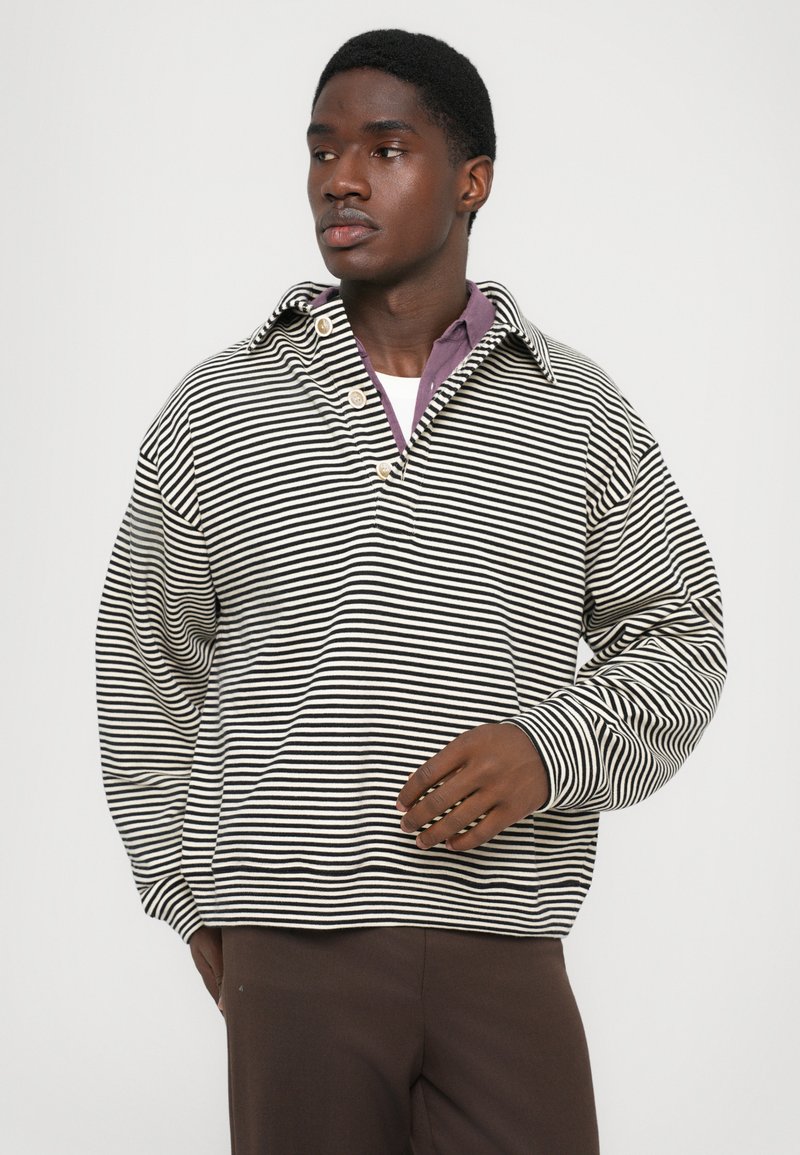 Man in een zwart-wit gestreepte lange mouwen shirt, gedragen over een paarse polo en met een bruine broek, kijkt naar links.