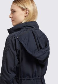 Giacca leggera navy con cappuccio, tessuto strutturato, vita elastica e tasche con zip laterali. Presenta un logo sul collo posteriore.