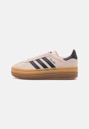 Sneaker Adidas Gazelle beige avec des bandes noires, un talon noir et une épaisse semelle en gomme superposée sur un fond blanc.
