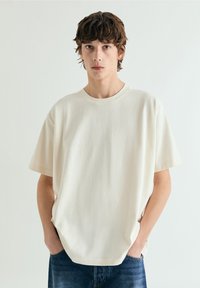 Hombre joven con cabello corto castaño, vistiendo una camiseta blanca lisa de gran tamaño y vaqueros azules, de pie con las manos en los bolsillos.