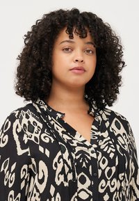 Blouse à manches longues noire et crème avec des motifs abstraits, dotée d'une fermeture à boutons, d'un col et d'un tissu texturé.