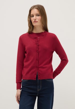 Woman with straight light brown hair wearing a buttoned red cardigan over a red top and dark blue jeans, standing with one hand on hip.