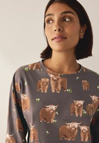 Graues Langarmshirt mit einem wiederholten Muster aus braunen Highland-Rindern und grünen Blumenakzenten, mit rundem Ausschnitt und weicher Textur.