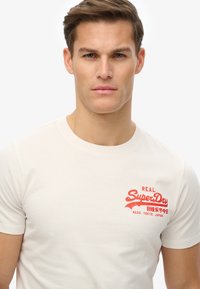 Camiseta de algodón color crema con mangas cortas. Presenta un logotipo gráfico rojo brillante en la zona izquierda del pecho, con texto en inglés y japonés.