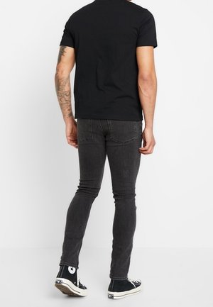 Černé tričko s krátkým rukávem spárované s tmavě šedými slim-fit džíny a černými vysokými teniskami; viditelné tetování na paži.