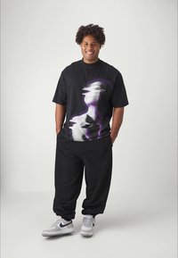 Junger Mann lächelt, die Hände in den Taschen, trägt ein schwarzes T-Shirt mit Grafik, lockere schwarze Hose und weiße Sneaker mit blauen Swoosh, steht vor weißem Hintergrund.