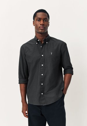 Jeune homme portant une chemise boutonnée gris foncé aux manches retroussées et un pantalon sombre, debout devant un fond clair uni.