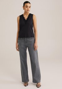 Črna brezrokavnik s čipkastim izrezom v obliki valov, v kombinaciji z visokim pasom in širokimi sivimi kavbojkami. Model stoji proti nevtralni ozadju.