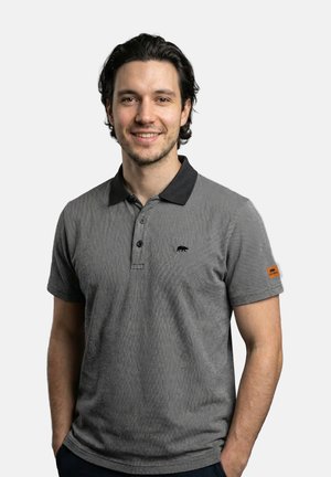 Lächelnder Mann mit dunklen Haaren, der ein graues Poloshirt mit kurzen Ärmeln, schwarzem Kragen und kleinen Logos auf Brust und Ärmel trägt. Hände in den Taschen.