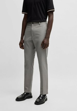 Man draagt lichtgrijze op maat gemaakte broek, zwarte leren loafers en een zwart shirt met korte mouwen met witte bies aan de mouwen.