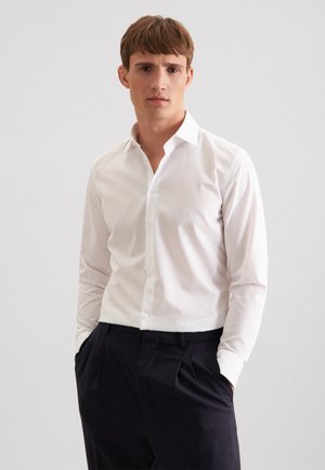 Jeune homme portant une chemise blanche ajustée à boutons et un pantalon bleu marine foncé, debout les mains dans les poches devant un fond uni.