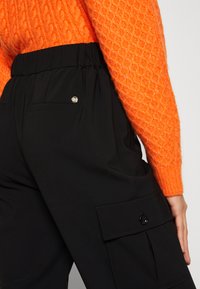 Sorte cargo bukser med sidelommer, elastisk talje og detaljer med sølv trykknap. Kombinert med en teksturert oransje genser.