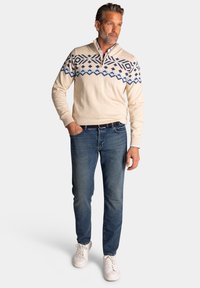 Beige gebreide trui met blauw geometrisch patroon, kwart-ritssluiting, en lange mouwen, gecombineerd met blauwe jeans en witte sneakers.