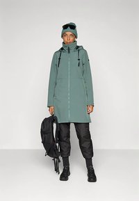 Długa, jasnozielona nieprzemakalna kurtka z wysokim kołnierzem, zestawiona z czarnymi ocieplanymi spodniami i czarnymi trampkami. Dodatki to czapka beanie i okulary przeciwsłoneczne.