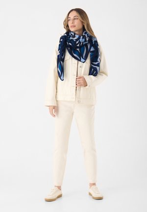 Giacca di jeans bianca sopra una maglietta crema, abbinata a jeans crema e sneakers beige. Una sciarpa a fantasia blu e navy aggiunge colore e texture.