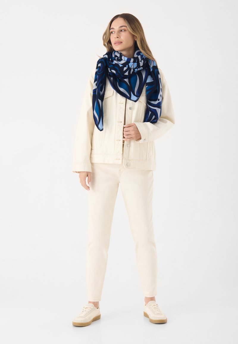 Giacca di jeans bianca sopra una maglietta crema, abbinata a jeans crema e sneakers beige. Una sciarpa a fantasia blu e navy aggiunge colore e texture.