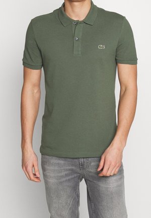 Homme portant un polo manches courtes vert olive avec un col et deux boutons, associé à un jean gris clair.