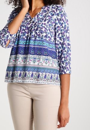 Kobieta ubrana w bluzkę z kwiatowym i paskowanym wzorem w odcieniach niebieskiego, fioletowego i białego, z rękawami do łokcia, oraz beżowe spodnie.