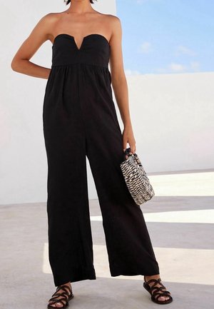 Musta olkaimeton haalari, jossa on V-aukko ja leveät lahkeet, yhdistettynä kudottuun mustavalkoiseen käsilaukkuun ja mustiin remmisandaaleihin.