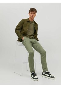 Veste en coton verte sur un t-shirt olive, associée à un pantalon vert clair et des baskets noires avec des accents blancs, assis sur un tabouret blanc.