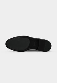 Chaussure habillée noire vue de dessous, montrant une semelle en caoutchouc texturée et un talon sur un fond clair et uni.