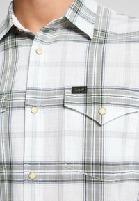 Camicia a quadri nei colori bianco, verde e blu. Tessuto in cotone con un motivo testurizzato, dotata di taschino sul petto e dettagli in metallo dorato.