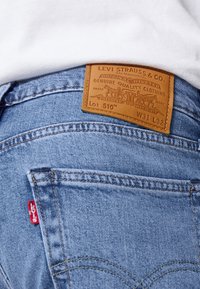 Kék farmernadrág barna bőr címkével, amelyen fényes szöveg és grafikák találhatók. Piros Levi's felvarró a hátsó zseben. Kényelmes szabás.