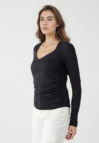 Chemise noire à manches longues avec un col en V, dotée de côtés froncés, en tissu lisse et extensible, associée à un pantalon de couleur claire.